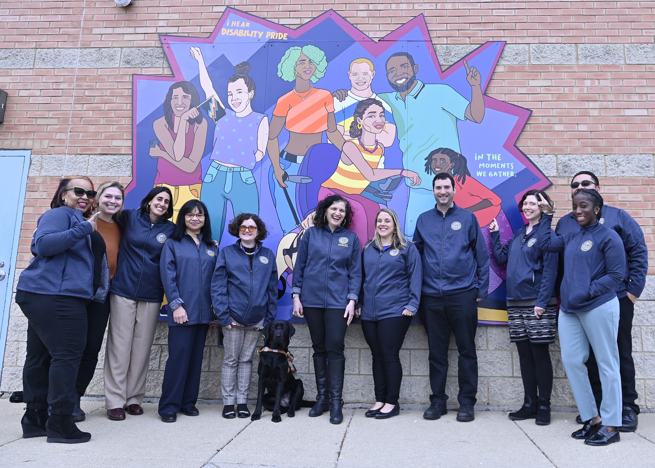 MOPD staff surround Commissioner Arfa outside the Central West Community Center building at a 2023 event. Most staff are wearing blue MOPD jackets. 